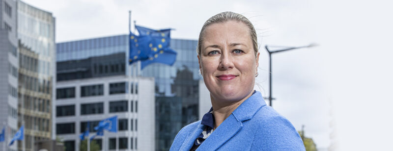 Jutta Urpilainen sinisessä jakkupuvussa Brysselissä, taustalla liehuu EU-lippuja.